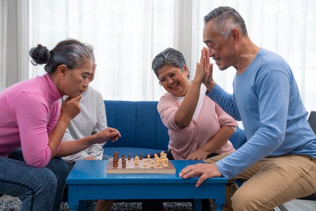 A family gathers around a chess table while one is thinking about the next move the other is high fiving a family member in celebration