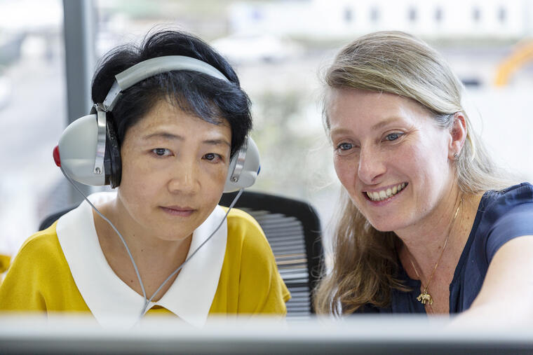 An audiologist guides a patient through a hearing test. They are both sitting and are looking at the computer screen. The patient is seen wearing a headset, while the audiologist is pointing tot the screen.